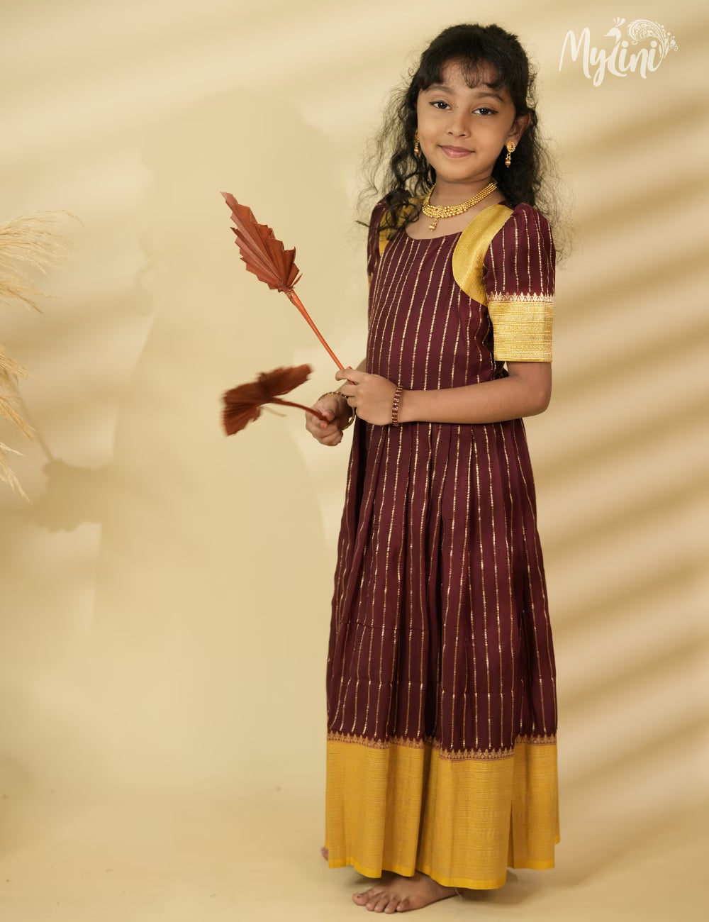 Young girl in a maroon and yellow traditional outfit holding a broom against a beige background