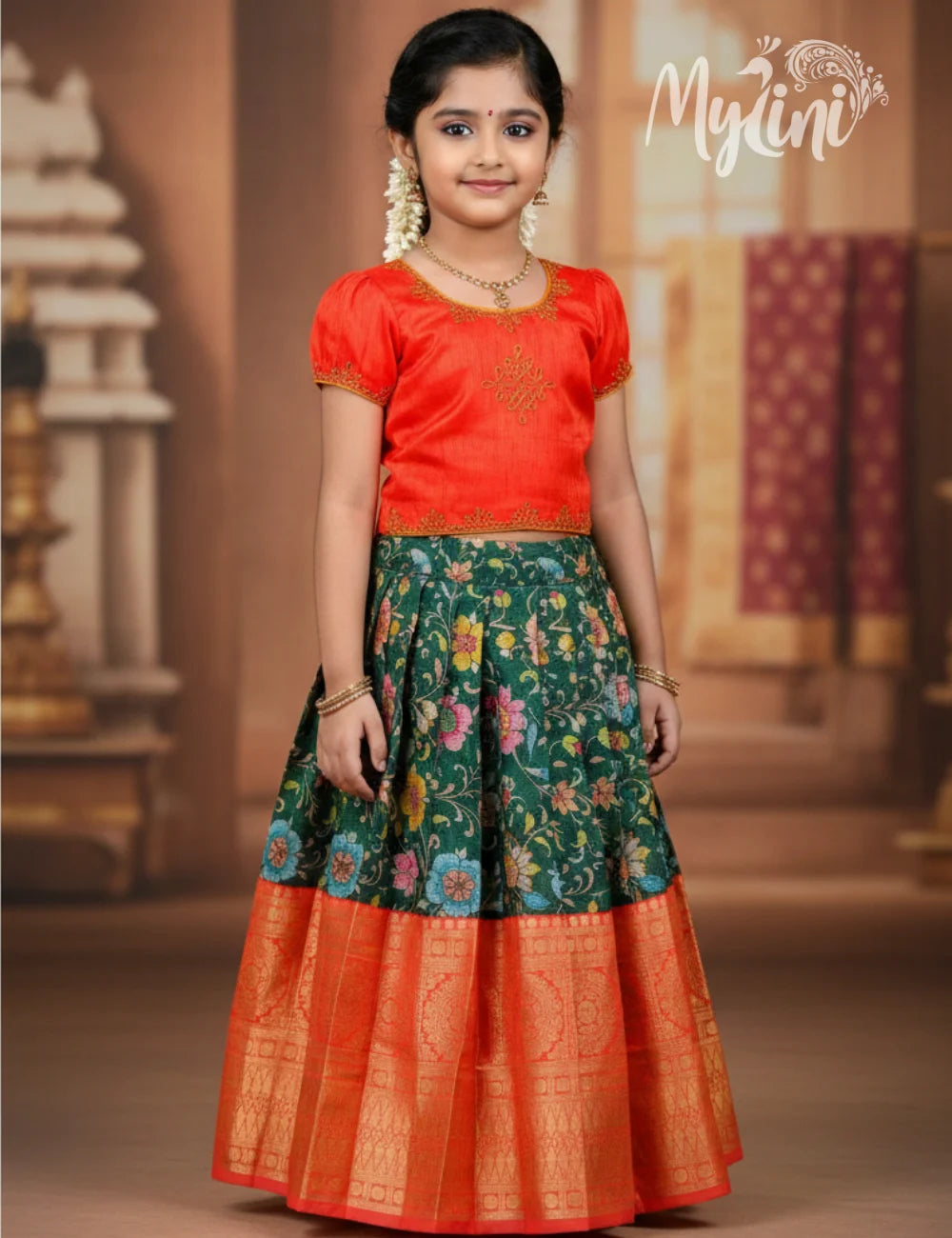 Young girl wearing a traditional outfit with a red top and floral skirt, standing in a decorated indoor setting.