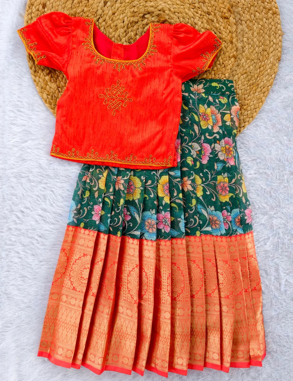 Young girl wearing a traditional outfit with a red top and floral skirt, standing in a decorated indoor setting.