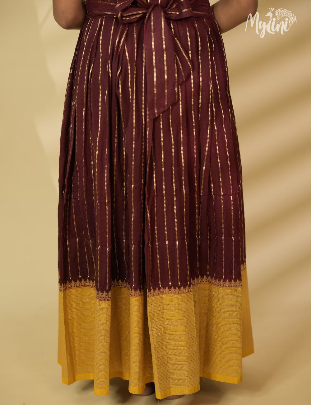 Young girl in a maroon and yellow traditional outfit holding a broom against a beige background